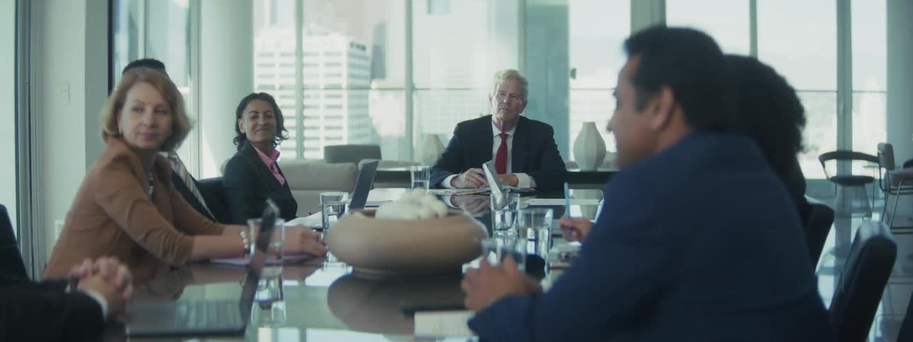 Many people seated around a glossy conference table in a modern bright office where an older man in the center is looking forward while two women and two men look towards the left foreground all framed by large glass windows