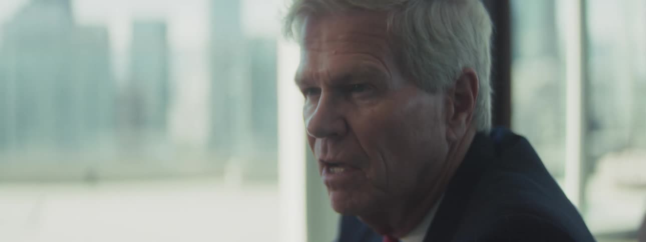 The video shows a closeup profile shot of an older man with gray hair appearing to be in a business or office setting possibly wearing a suit he is seated next to a large window with a blurry view of a cityscape or buildings outside the man is speaki