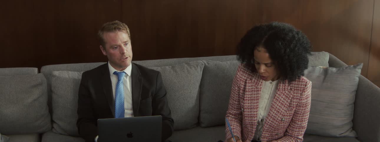 A man in a dark suit gestures while explaining information displayed on a laptop to a woman in a tweed jacket who intently listens and takes notes while they sit on a gray couch in a professional lounge setting