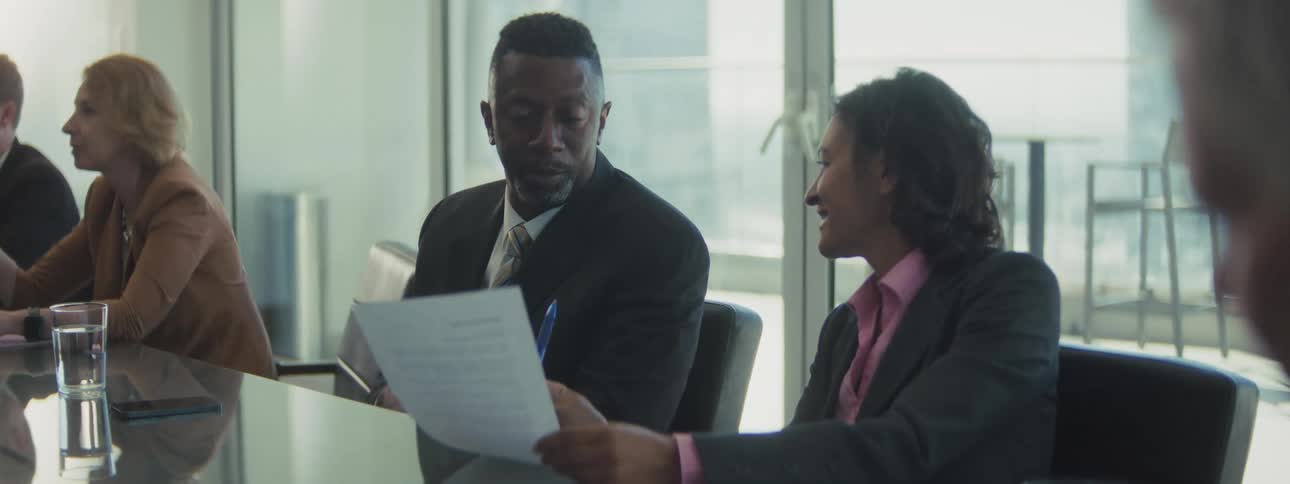 A man in a suit is sitting next to a woman in a bright conference room looking intently at a document she is holding they are engaged in a serious discussion with the man speaking and gesturing as he reviews the paper