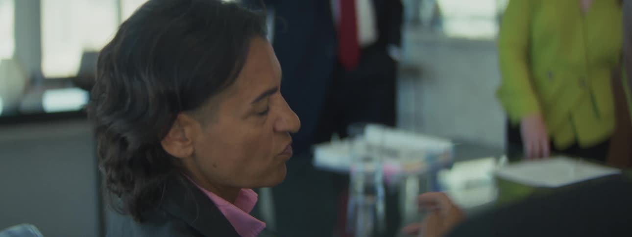 A closeup sideprofile shot features a woman in business attire actively speaking and gesturing with a pen at a reflective conference table fully engaged in a serious discussion in a bright modern office while two other people stand on the back