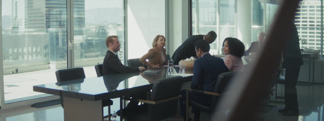 A modern sunlit conference room features a group of diverse professionals around a long reflective black table with a bowl of objects at the center as one black man in a suit speaks on a smartphone while others are seated or standing