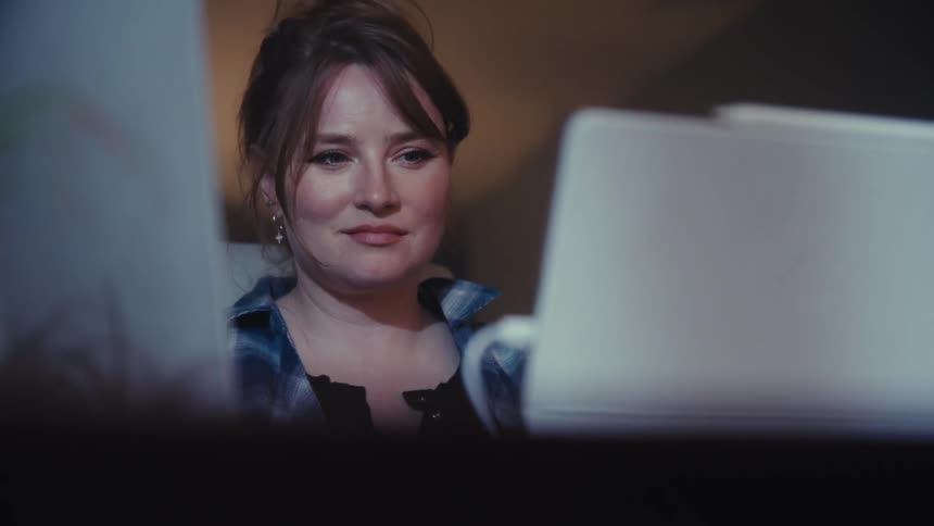 A woman with a plaid shirt is framed between two laptop screens in a dimly lit room showing a pensive expression as she concentrates on her work occasionally resting her hand on her chin