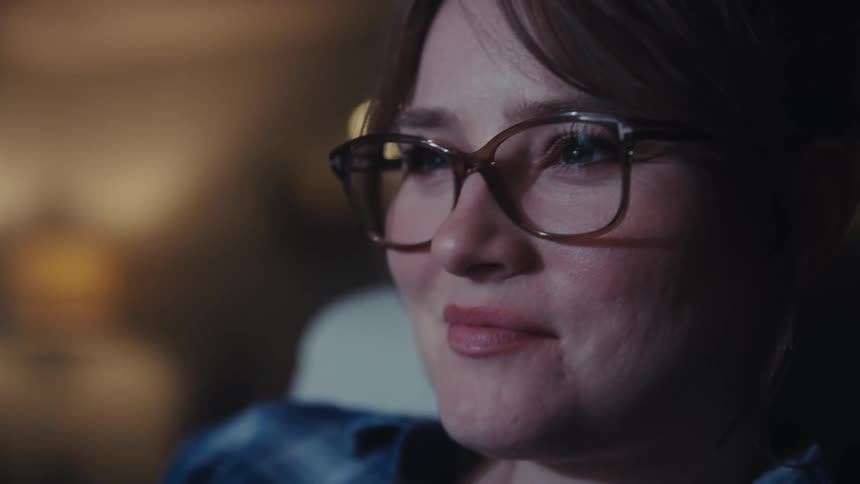 In a cozy dimly lit room a woman with glasses smiles warmly while watching something the camera gently moving closer to her content face