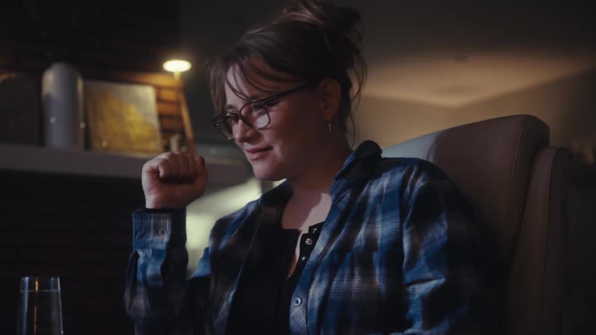 A woman with her hair in a bun and wearing glasses and a blue plaid shirt is sitting and looking at a screen she smiles appears to be enjoying what she sees and rests her chin on her hand