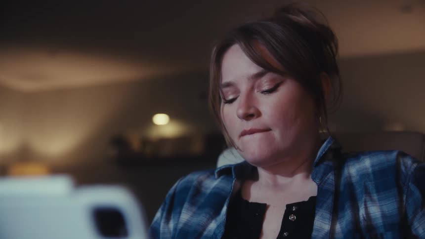 A woman in a blue plaid shirt is shown wiping her eyes as if she has been crying or is very tired she looks down and focuses her attention on something offscreen possibly a computer or document with a concerned expression