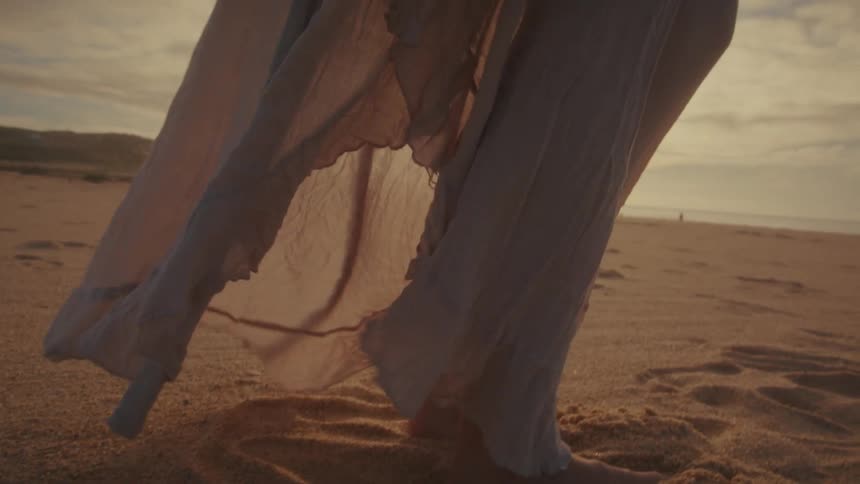 The lower portion of a light flowing garmentis blowing in a gentle breeze against the backdrop of a vast sunny and slightly hazy beach with visible sand dunes