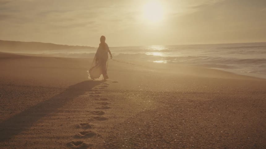 A woman wrapped in flowing fabric walks away from the camera on a wide sandy beach toward the ocean silhouetted against the bright golden light of a sunset