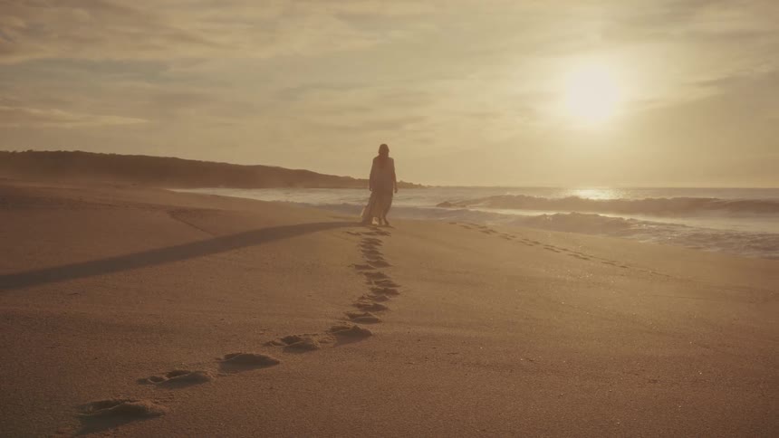 A solitary woman walks away along the golden sandy beach towards the bright setting sun leaving a trail of deep footprints behind her as gentle waves lap at the shore