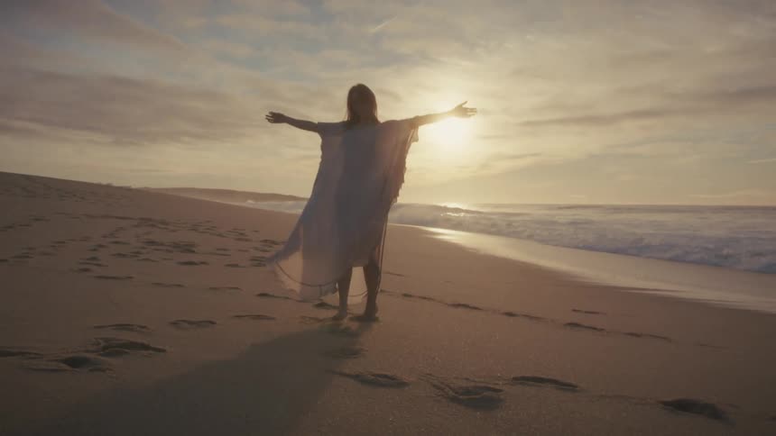 A young woman walks barefoot on the beach sand at sunset excited with the setting sun behind her