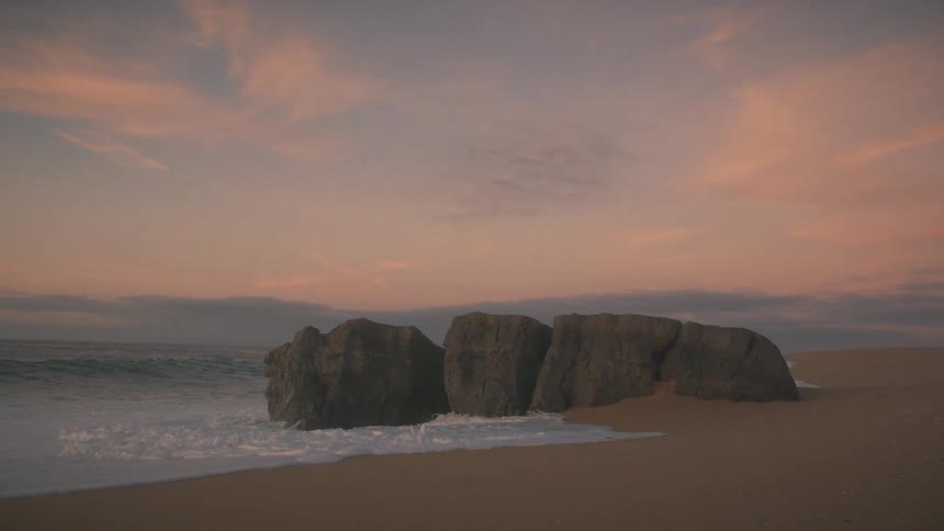 Massive ocean waves crash forcefully against a large dark rock formation on the sandy shoreline surging white foam around the stone base beneath a soft pastel pink sunset sky