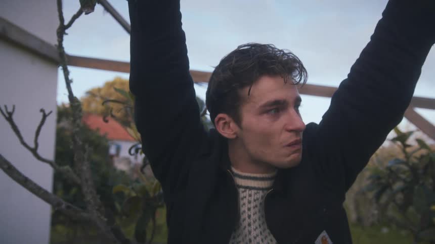 A young man stands outdoors behind a garden holding a platform and wearing a jacket his eyes filled with sadness at midday