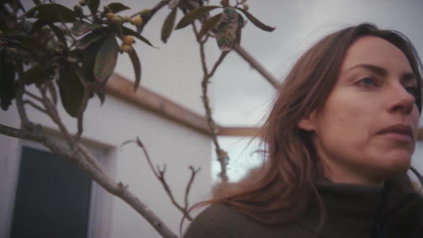 A closeup shot of a woman with an intense expression adjusting her collar is framed by the branches of a loquat tree with small fruits under an overcast sky then she looks up thoughtfully