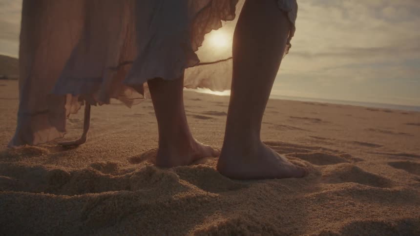 Barefoot woman stands on the beach as her long dress moves in the breeze