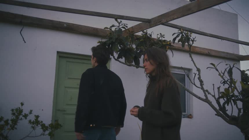 A man and a woman stand outside a white building beneath a wooden structure during twilight engaged in a conversation using expressive hand gestures before briefly turning away
