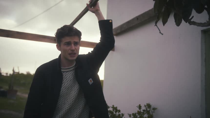 A distressed young man holds onto a wooden beam while speaking intensely to a woman outdoors