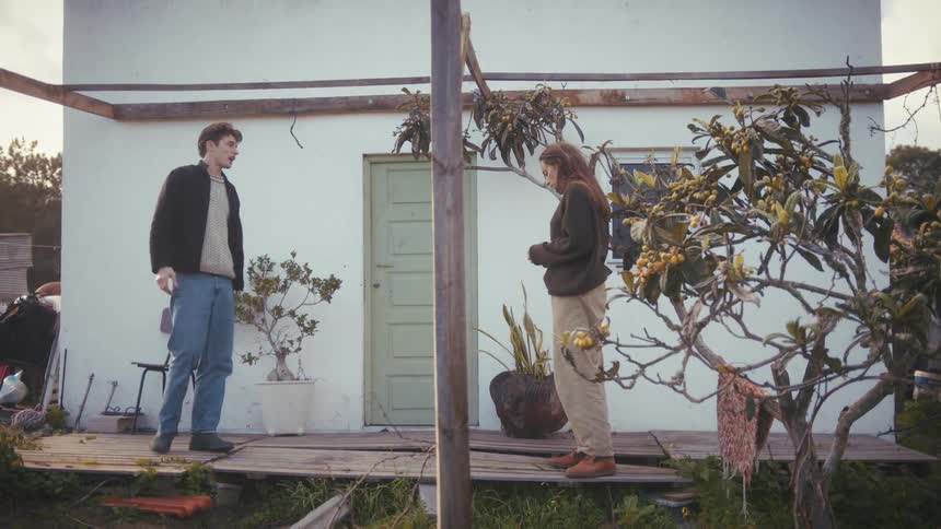 Man gestures while talking to another person on a wooden porch in front of a house
