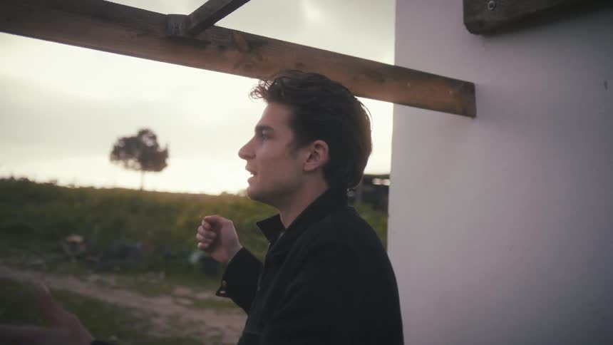 A young man wearing a black jacket and patterned sweater sits outdoors speaking passionately with animated hand gestures before standing up to lean against and touch the wooden beam above him in a rural setting
