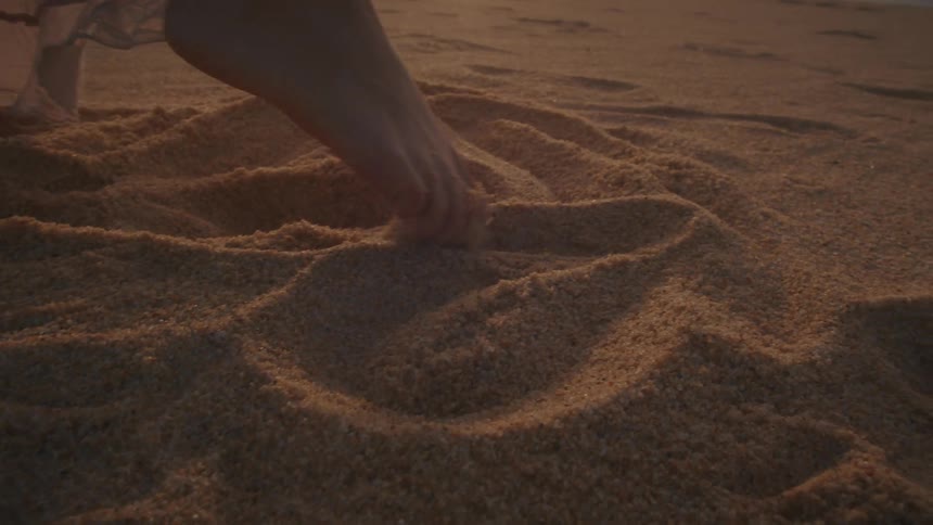 Woman stands on the beach making designs in the sand with her bare foot