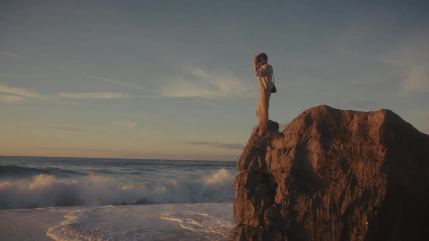 A man stands atop a large coastal rock at sunset holding a camera up to his face to capture the dramatic ocean waves breaking below and the light over the water