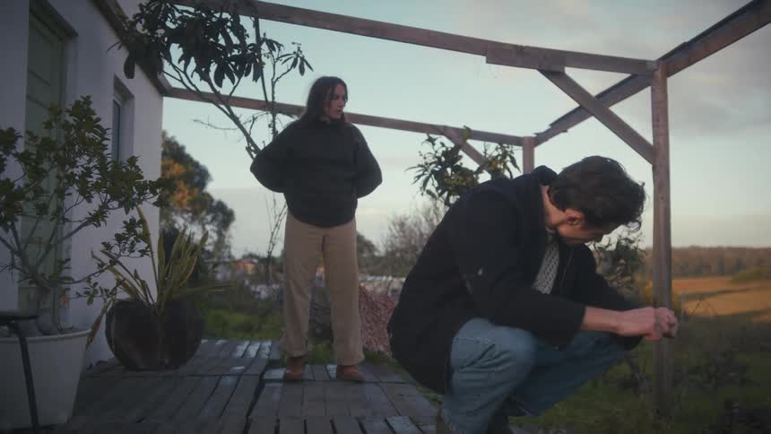 Aman crouches outside on a wooden deck while a woman stands behind him looking out onto a rural landscape at dusk