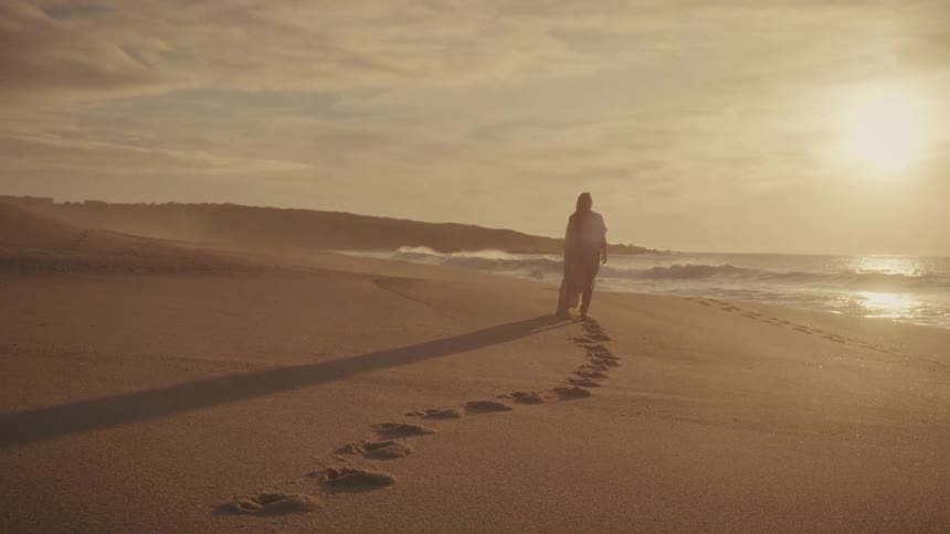 Footprints in the sand lead to a woman walking on the beach as the sun shines brightly through the clouds