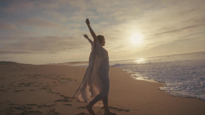 Woman walking on the sand with arms raised near the water at sunset
