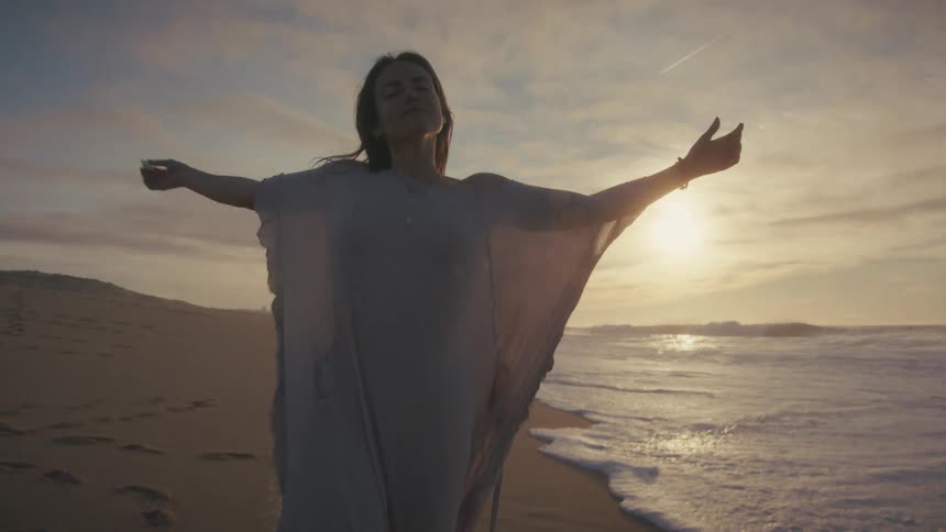On a windswept beach at sunset a woman in a long flowing dress turns towards the light and slowly raises her arms to the sky embracing the vastness of the ocean and the golden air