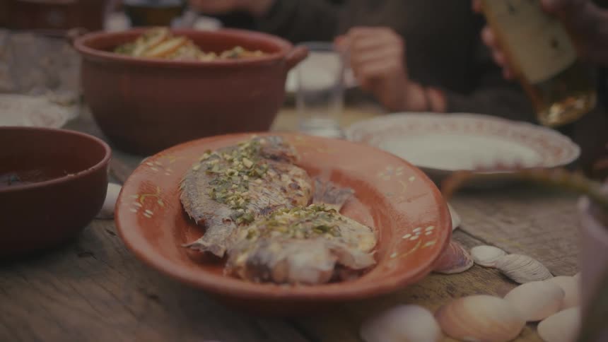 A closeup shot focuses on a platter of whole cooked fish garnished with herbs placed on a rustic table amidst bowls of food while a hand pours a beverage into a glass during a gathering