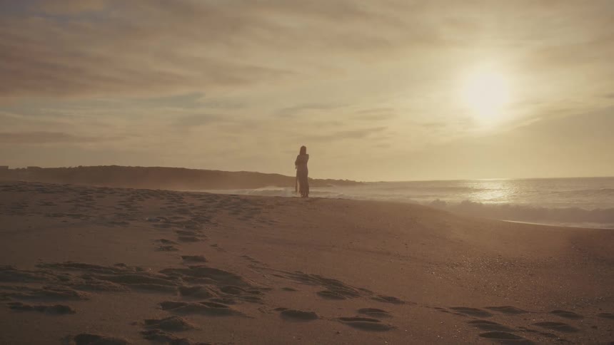 A wide shot on a vast sandy beach at sunset shows the silhouetted figure of a woman standing near the waters edge gazing out at the bright sun and the waves breaking in the golden light