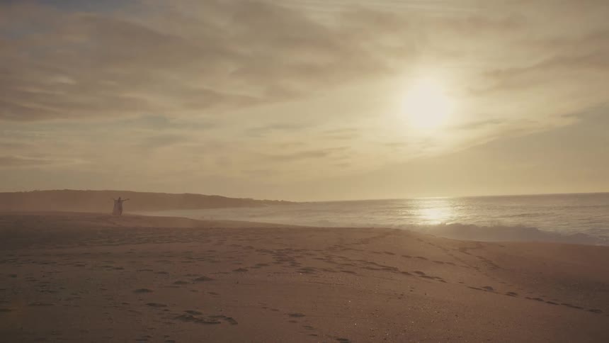 Woman walks on the beach as the sun glows through the clouds