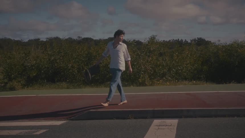 A barefoot man walks casually along a red paved path next to a road surrounded by greenery and a partly cloudy sky