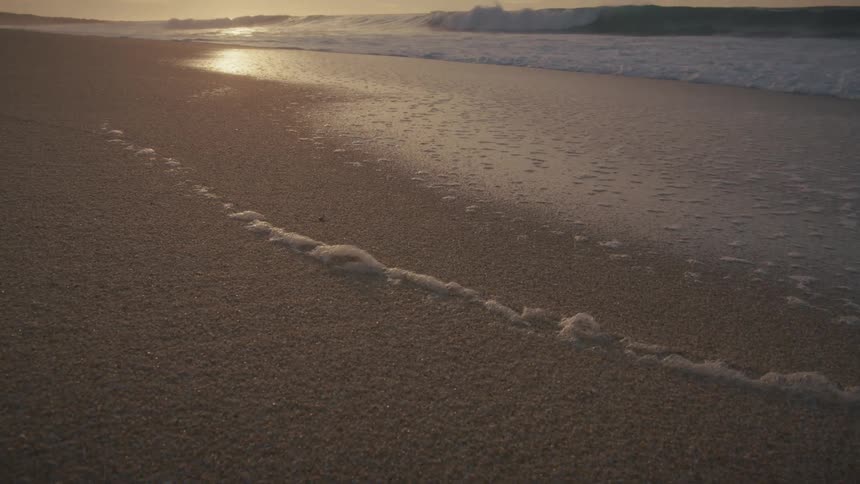 Waves gently roll onto the sand leaving a frothy trail as the sun sets behind them