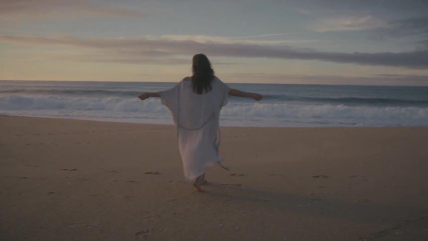 A woman in a long dress walks barefoot on the beach sand at sunset