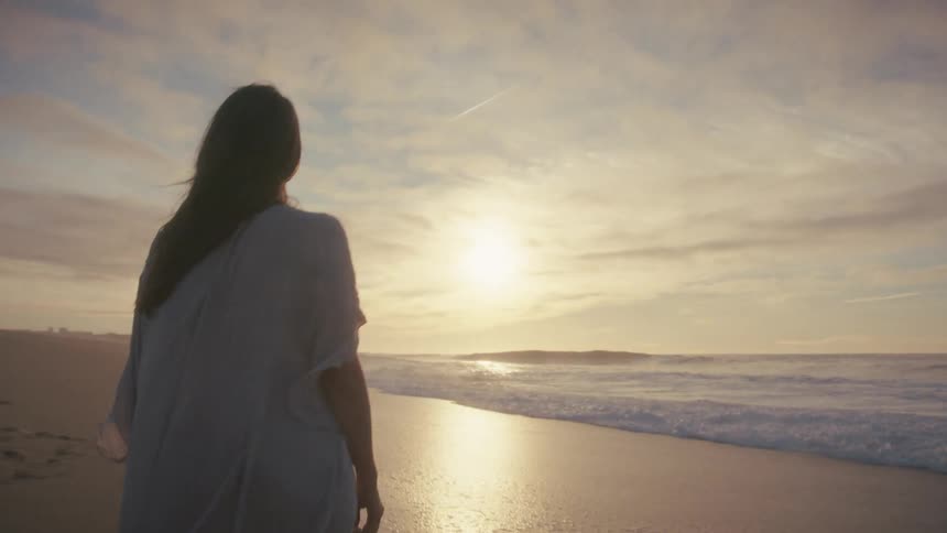 A woman in a light top walks on a sandy beach during a dramatic sunset first raising her arms toward the sky and then strolling near the waves looking out at the ocean as the wind blows her hair