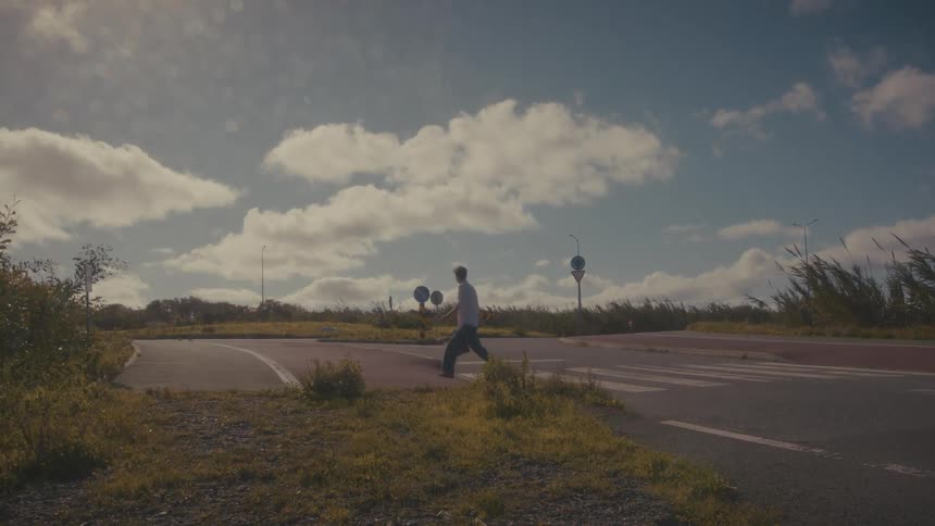 A man wearing dark clothes is walking across a small paved road at an intersection with overgrown grass and bushes surrounding the area under a bright cloudfilled blue sky
