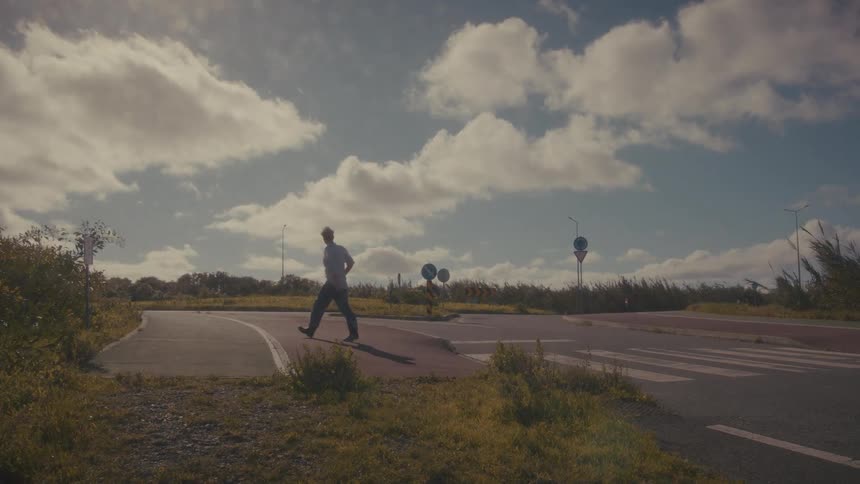 Under a wide blue sky filled with bright cumulus clouds a man casually walks across a marked crosswalk and continues along a curving empty road surrounded by low grass