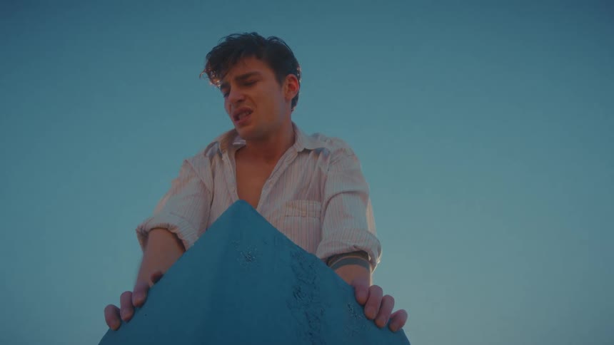 Man leaning on a pointed blue object under a blue sky frowns and grimaces upset