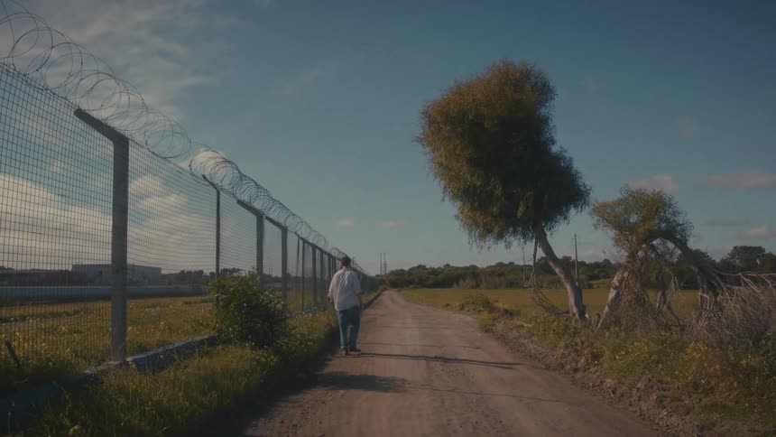 Person walking beside metal fence with barbed wire under clear sky