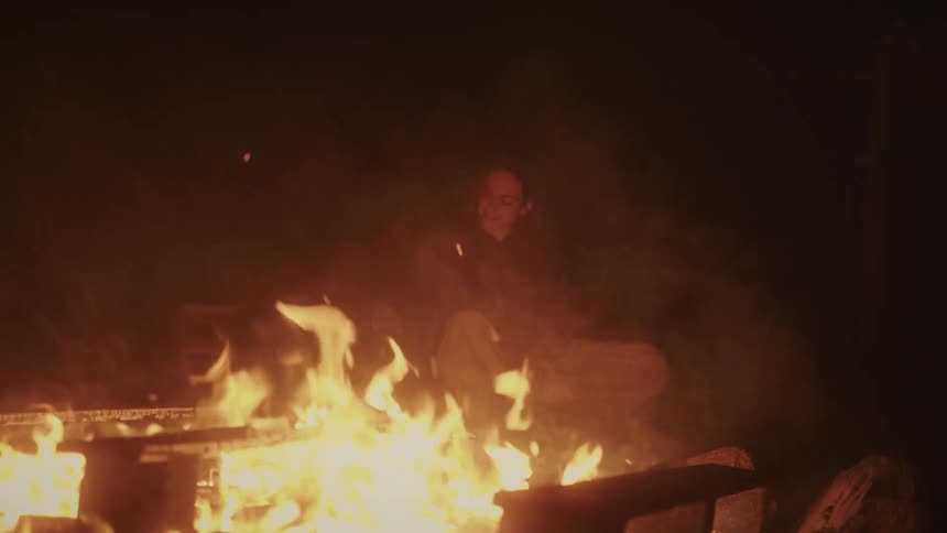 A woman smiles warmly as she gently pets her dog while sitting cozy next to a crackling campfire