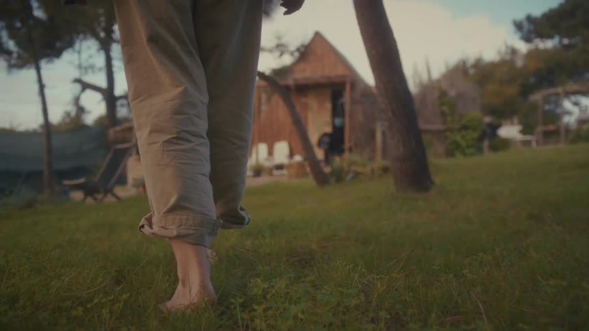 A person wearing loose linen trousers strolls barefoot across a lush lawn playfully skipping as they move through a tranquil garden toward a rustic wooden cabin shaded by tall pine trees