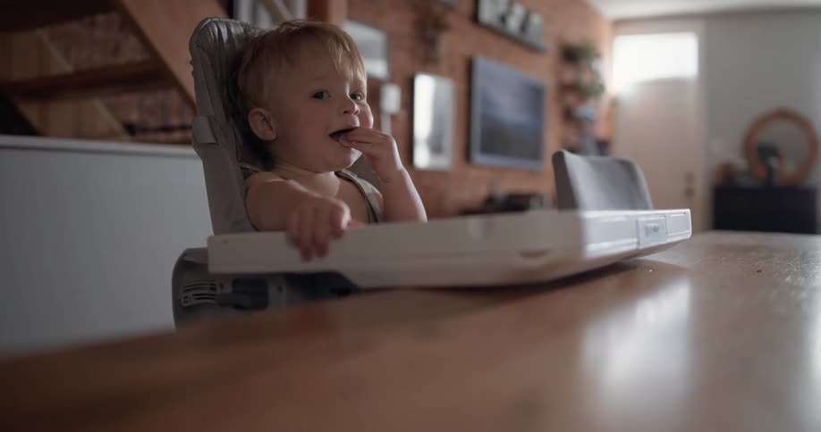 A toddler eats a snack in a high chair at a wooden table in a softly lit home