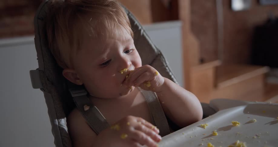 A messy toddler sits in a high chair happily munching on shredded bits of food thats spread across the tray