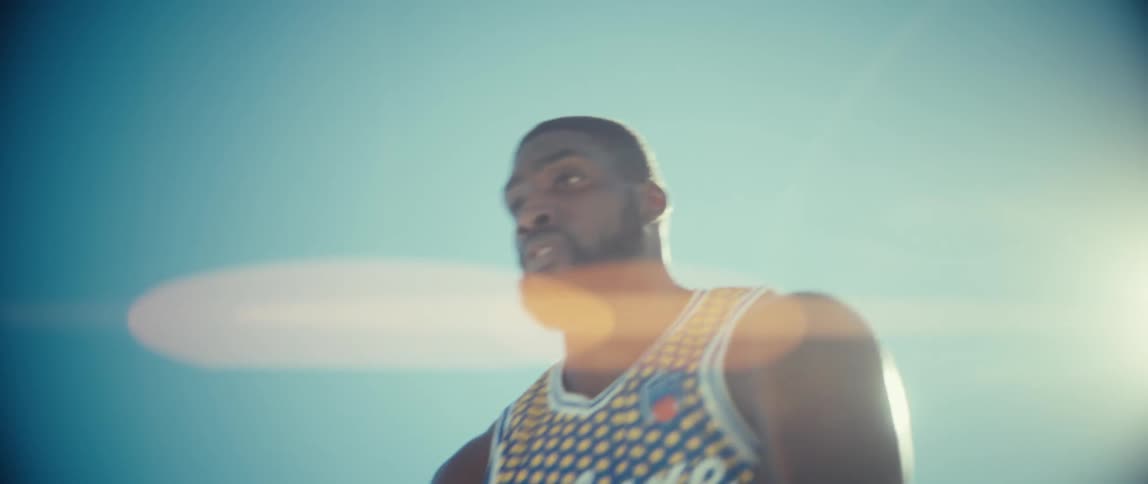 A basketball player holds a ball against a bright blue sky with a lens flare