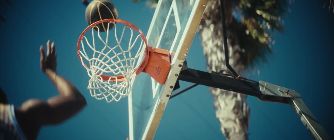 A basketball swishes through an outdoor hoop against a backdrop of blue sky and a palm tree