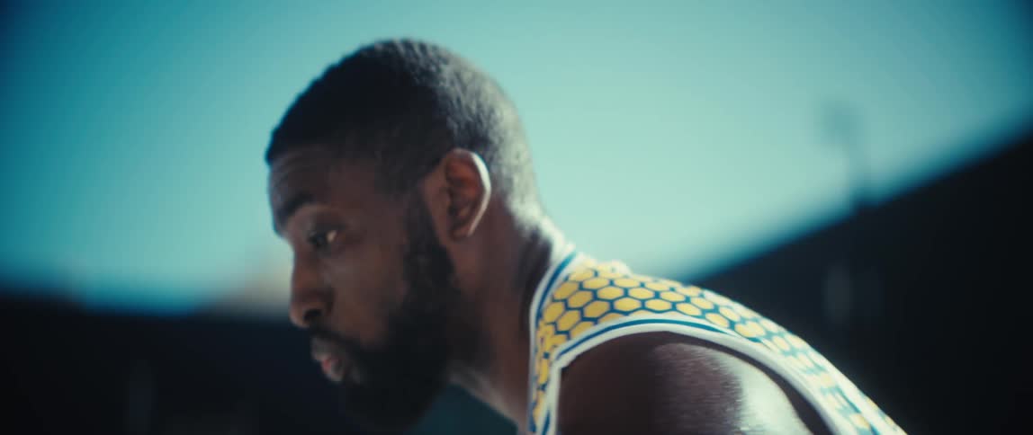 A closeup of a basketball player wearing a blue and yellow jersey shows his hands handling the ball and his intense focused expression against a bright blue sky