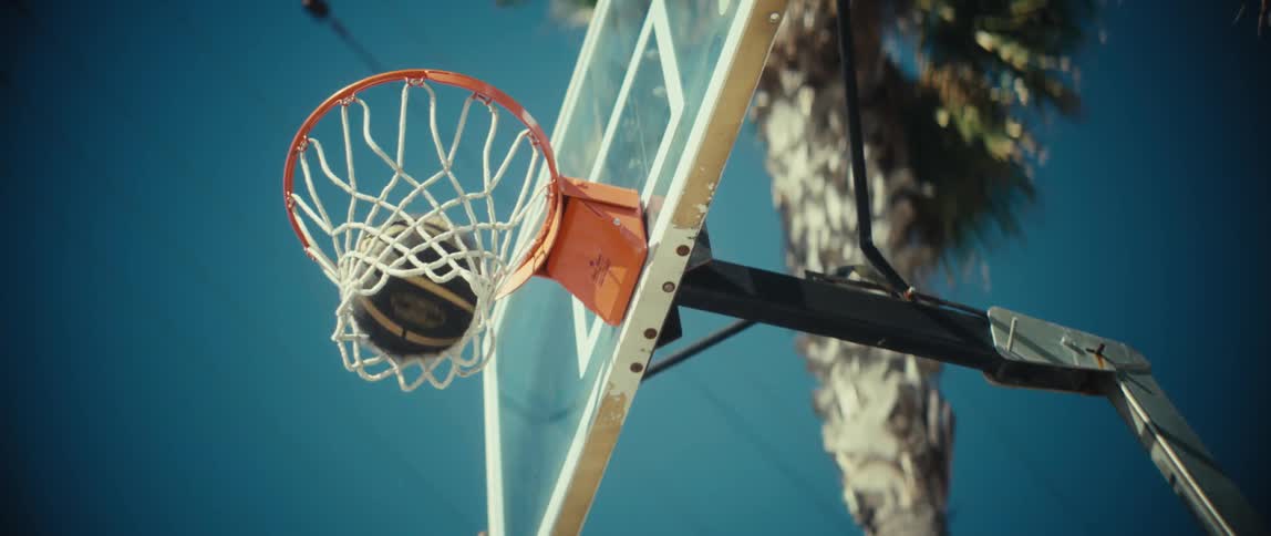 A young black man scores a basketball into a rim in an outdoor court during daytime