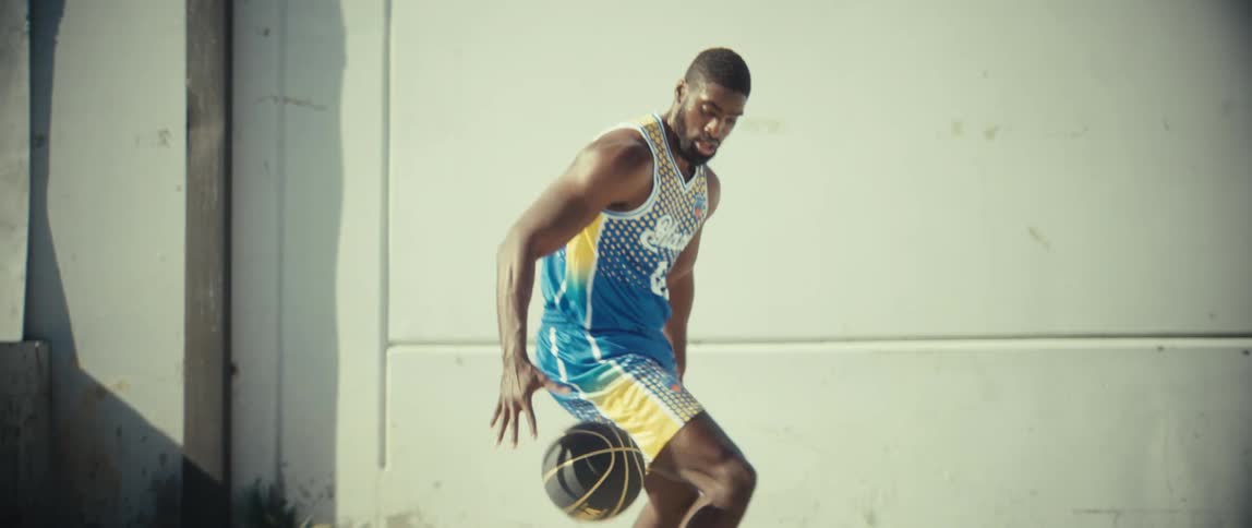 A young black male in sneakers and activewear plays basketball alone under the midday sun