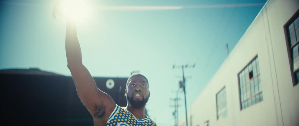 A young male athlete dunks the basketball into the net under the sun wearing a yellow jersey and excited after the play