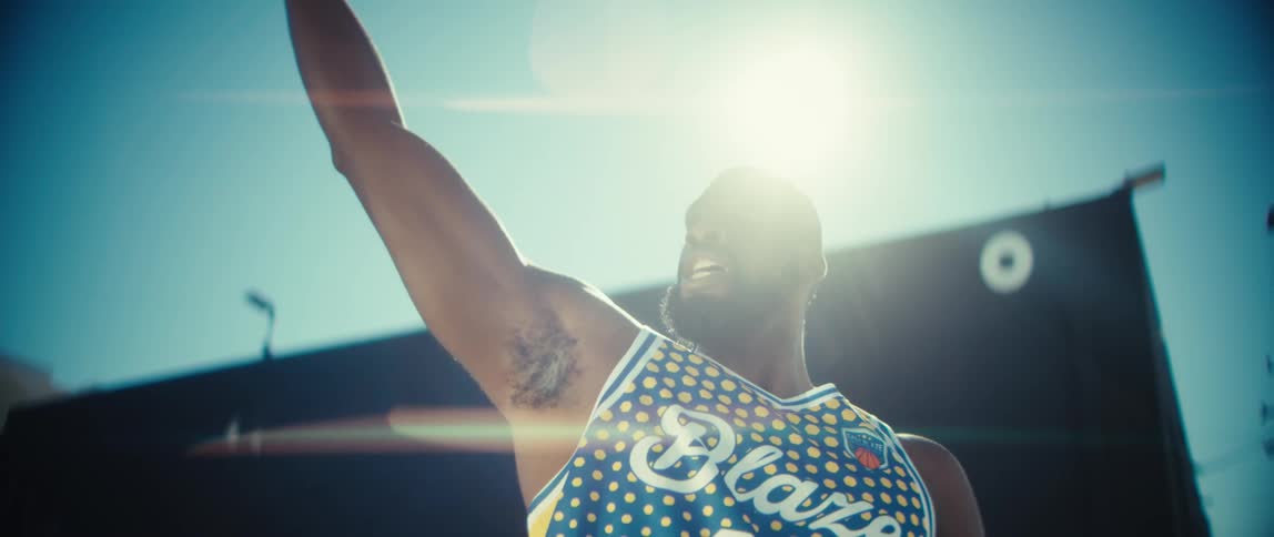 A basketball player dribbles shoots and celebrates a basket on a sunny outdoor court before bending over to catch his breath